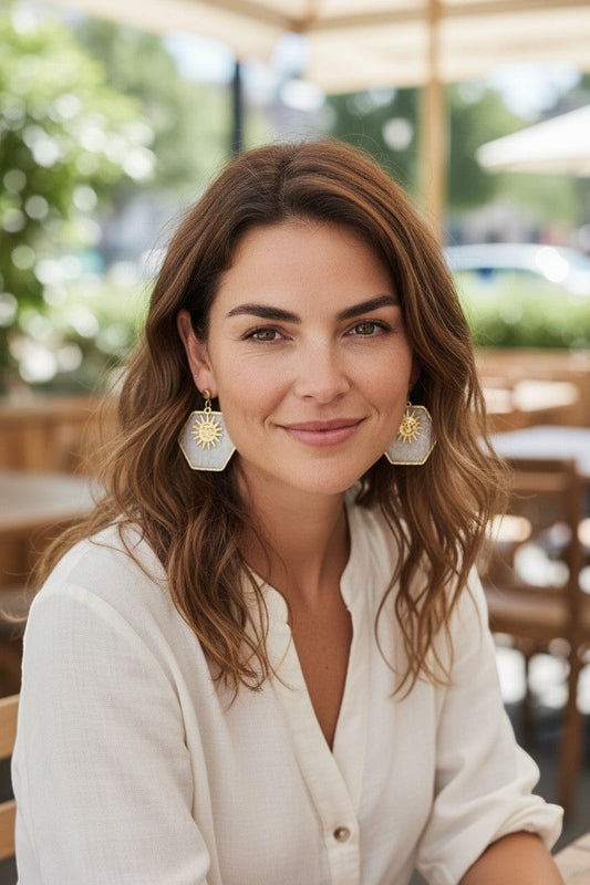 Lakelynns Designs white pearlescent hexagon earrings with gold sun charms and hooks. Model wears them with a white linen shirt.