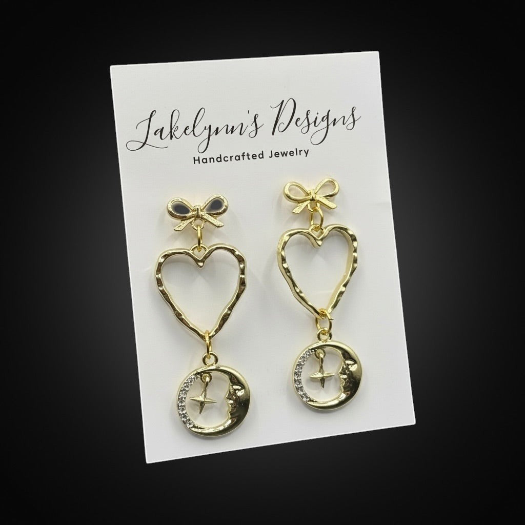 Lakelynns Designs gold-tone celestial heart and moon dangle earrings with bow accent and crystal details.
