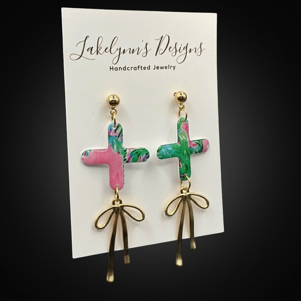Lakelynns Designs pink, green, and blue cross dangle earrings with gold bow charms.