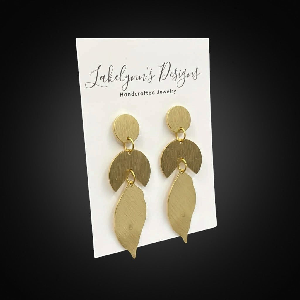 Lakelynns Designs geometric leaf dangle earrings in brushed brass-toned metal. Abstract design with circle, crescent, and leaf shapes.