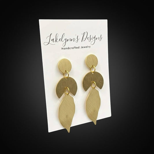 Lakelynns Designs geometric leaf dangle earrings in brushed brass-toned metal. Abstract design with circle, crescent, and leaf shapes.
