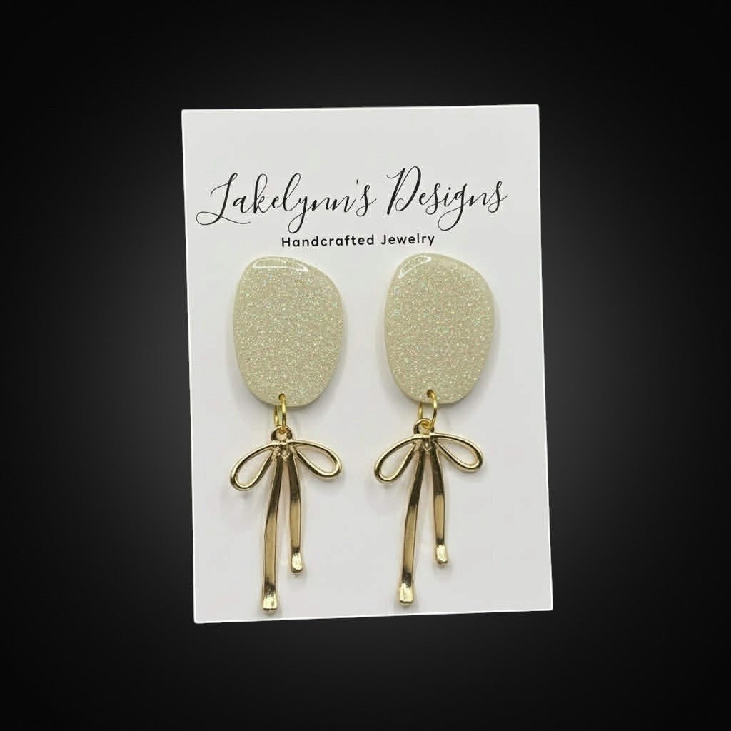 Lakelynns Designs glitter oval dangle earrings with gold-tone bow charms. Light champagne color with subtle sparkle.