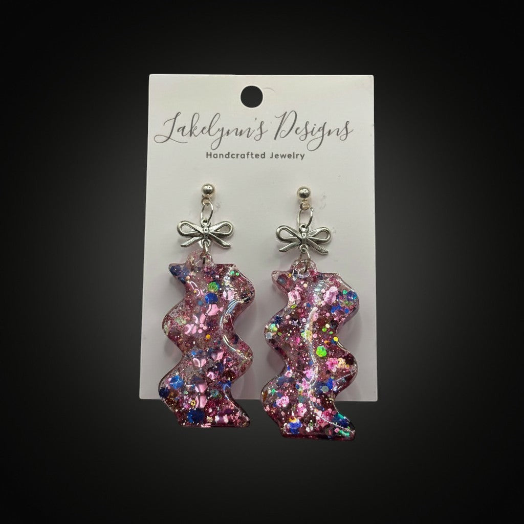 Lakelynns Designs glittery zigzag earrings with pink, purple, and blue iridescent glitter and silver-tone bow accents. Resin.
