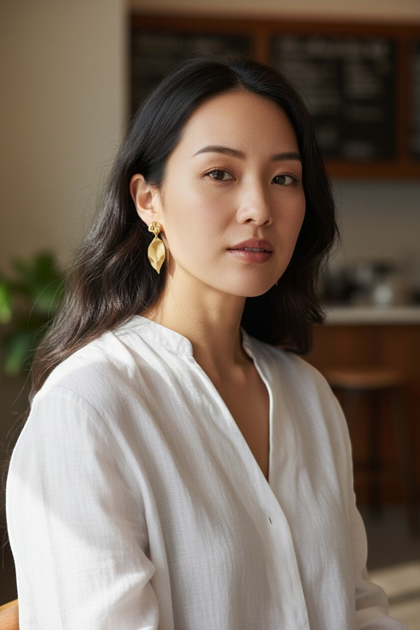 Lakelynns Designs gold-tone knotted leaf dangle earrings with brushed finish. Model wears white linen shirt.