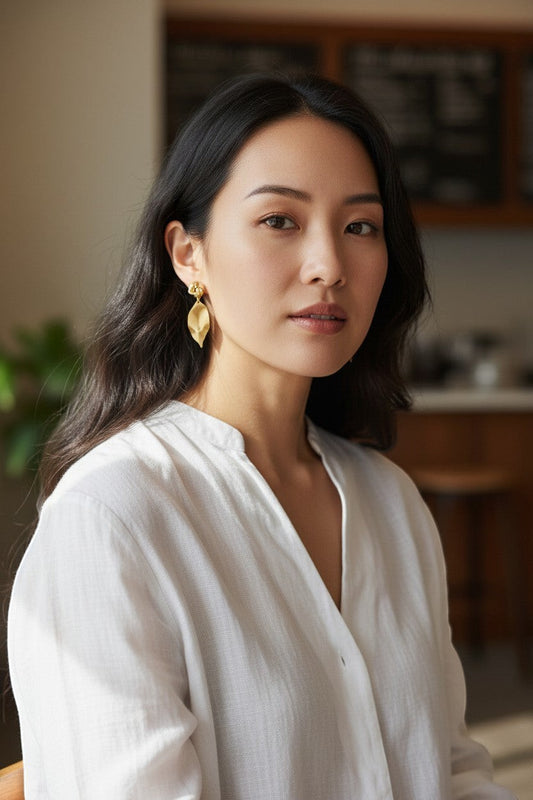 Lakelynns Designs gold-tone knotted leaf dangle earrings with brushed finish. Model wears white linen shirt.