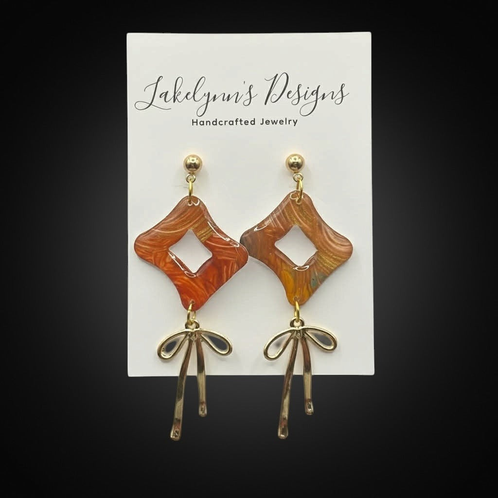 Lakelynns Designs rhombus dangle earrings with abstract marbled pattern in burnt orange, red, yellow, teal, and gold. Gold-tone bow charms dangle below.