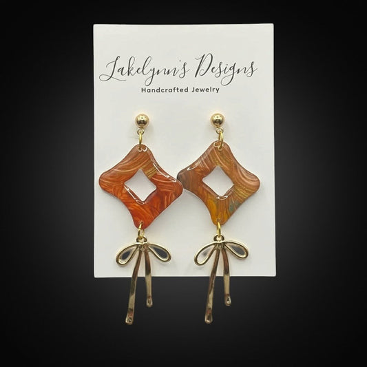 Lakelynns Designs rhombus dangle earrings with abstract marbled pattern in burnt orange, red, yellow, teal, and gold. Gold-tone bow charms dangle below.